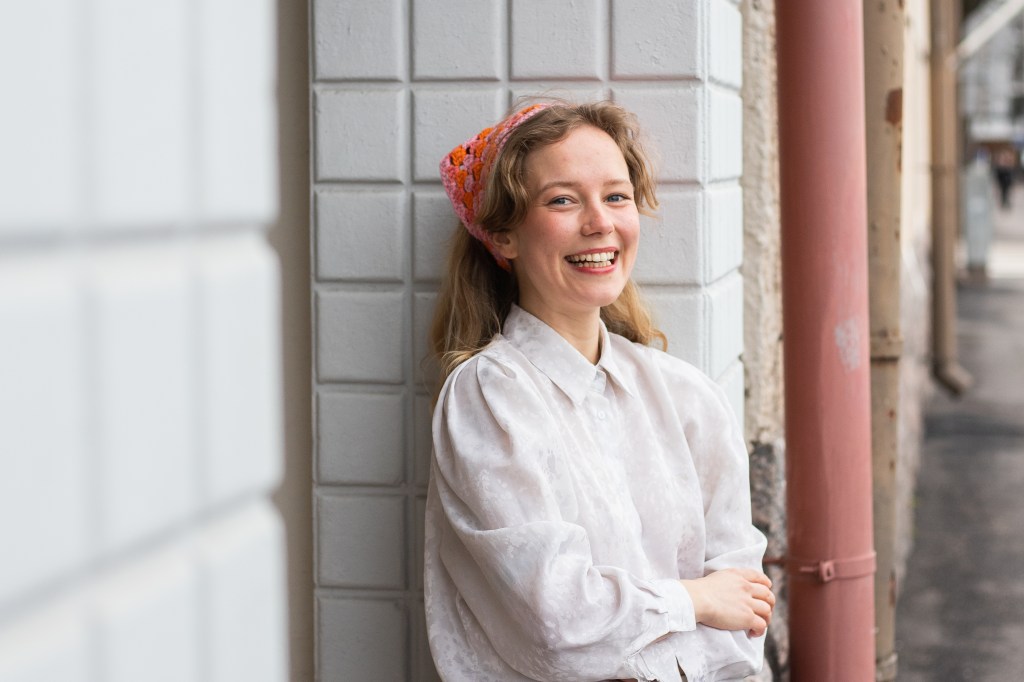 Lotta hymyilee ja katsoo kameraan ja nojaa valkotiiliseen seinään. Hänellä on päässään virkattu huivi ja yllään valkoinen kauluspaita.