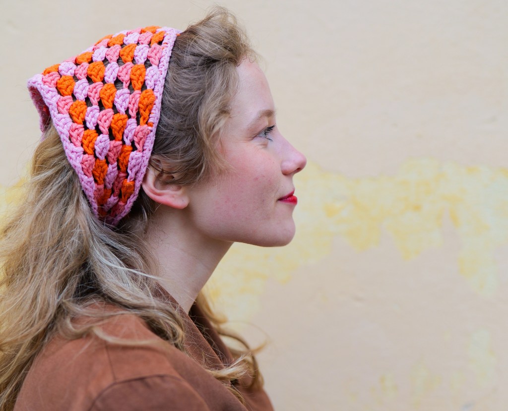 Kuvassa ovat Lotan kasvot sivusta päin kuvattuna. Hänellä on virkattu huivi päässään, punaista huulipunaa ja ruskea jakku. Takana on keltainen kiviseinä.