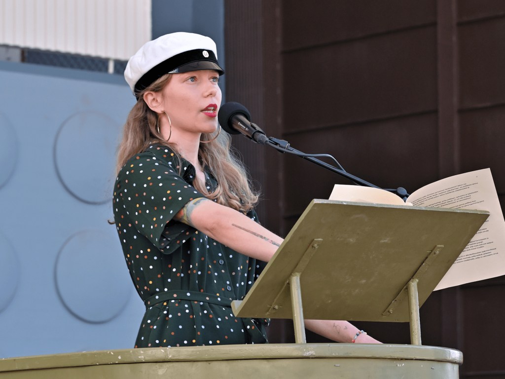 Lotalla on yllään pilkullinen, vihreä jumpsuit ja päässään ylioppilaslakki ja korvissaan isot renkaat. Hän puhuu mikrofoniin puhujankorokkeella Jyväskylän Lounaispuistossa.