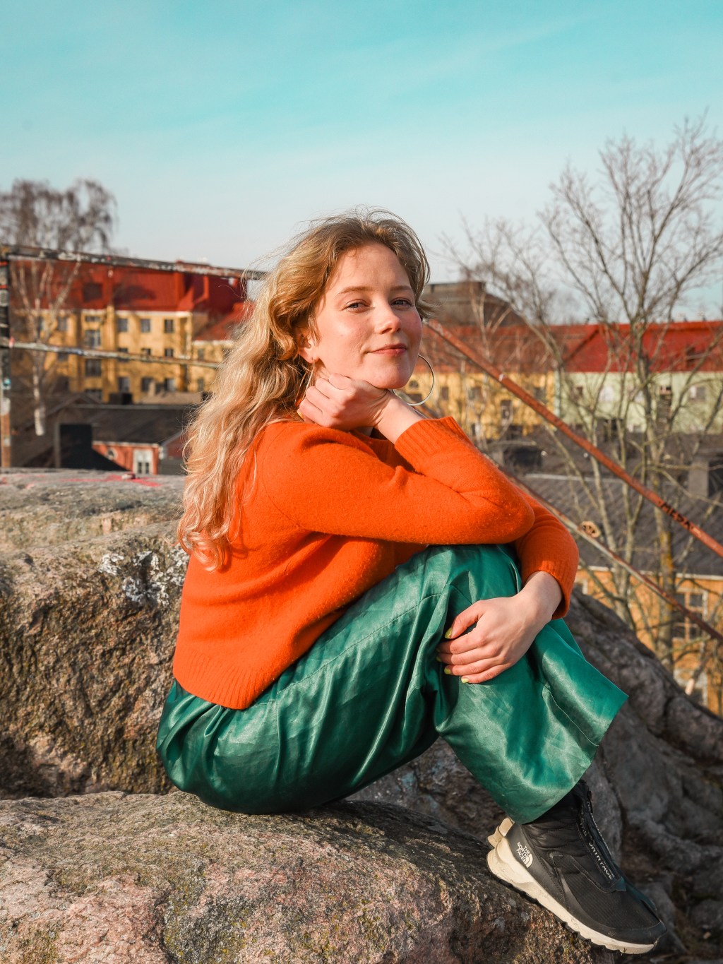 Lotta istuu kalliolla Helsingin Vallilassa ja nojaa polviinsa. Hänellä on päällä oranssi paita ja vihreät, kiiltävät housut. Takana näkyy taloja.