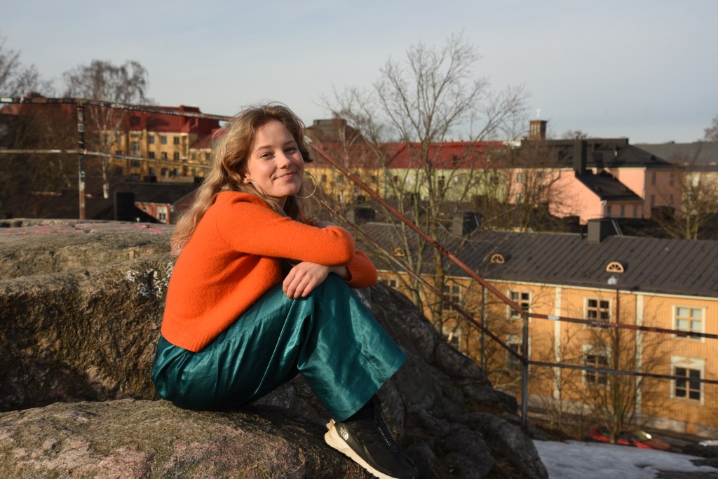 Lotta istuu kalliolla Helsingin Vallilassa ja nojaa polviinsa. Hänellä on päällä oranssi paita ja vihreät, kiiltävät housut. Takana näkyy taloja.