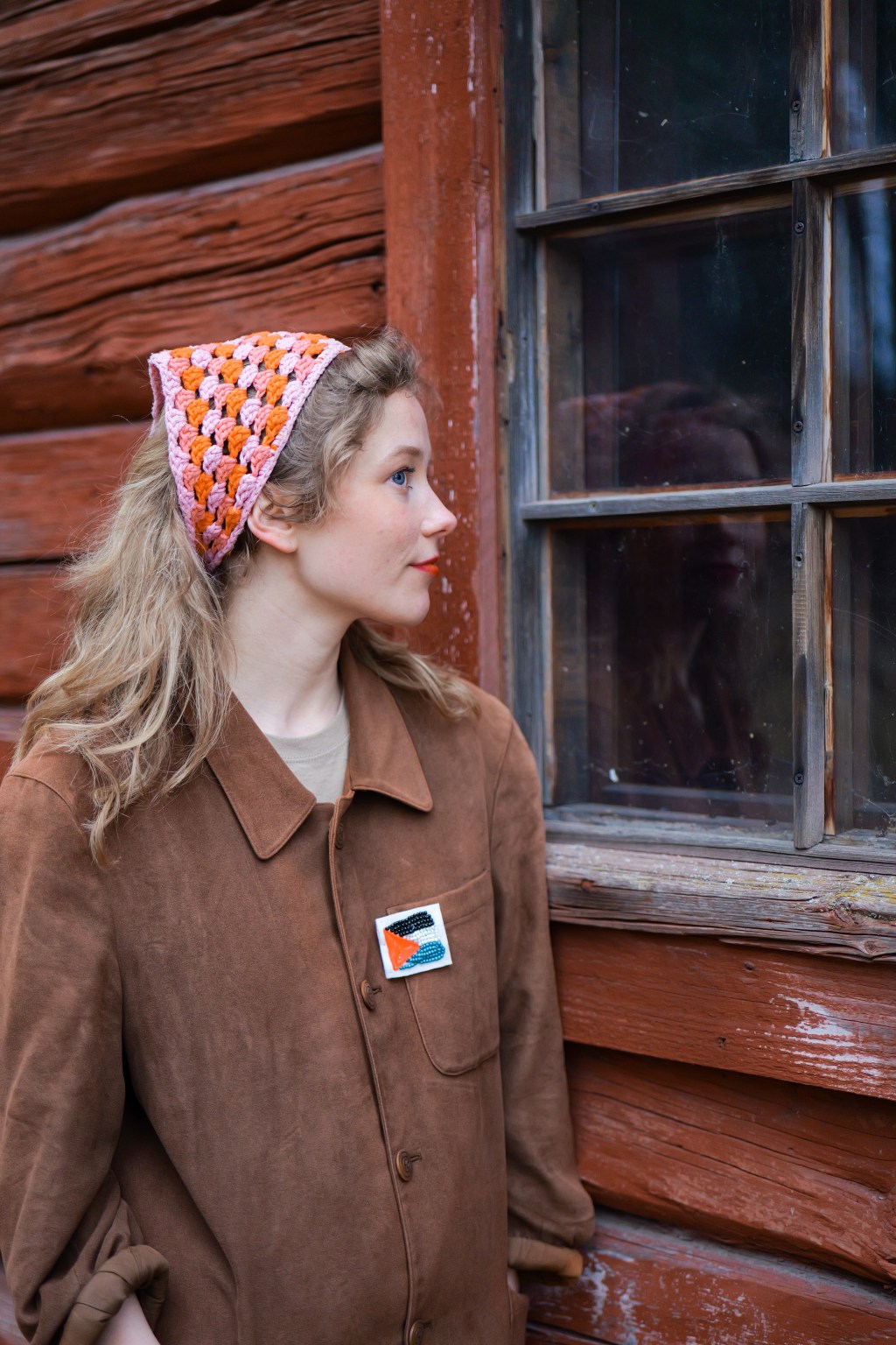 Kuvassa Lotta katsoo punaisen hirsituvan ikkunasta sisään. Hänellä on päässään virkattu huivi ja yllään ruskea takki, jossa on Palestiinan lippua muistuttava pinssi.