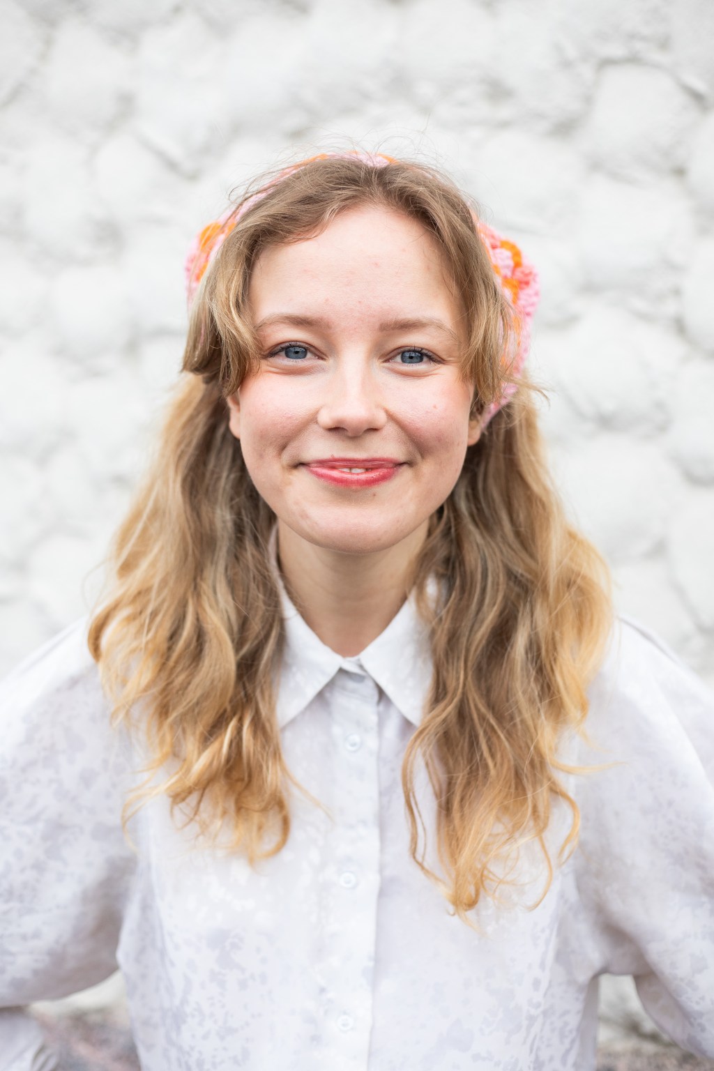 Lotta seisoo valkoisen kiviseinän edessä ja katsoo suoraan kameraan hymyillen. Hänellä on yllään valkoinen kauluspaita ja hiuksissaan virkattu huivi. Hiukset ovat edessä avoimina.