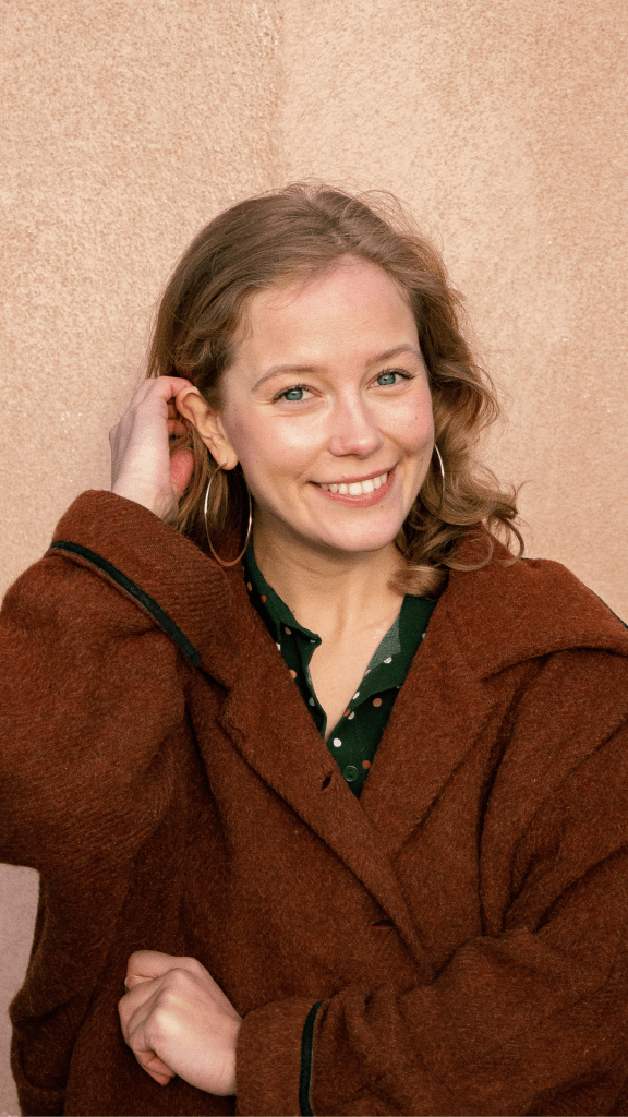 Lotta seisoo yksiväristä kiviseinää vasten, hymyilee kameraan ja sukii hiuksia korvan taakse.