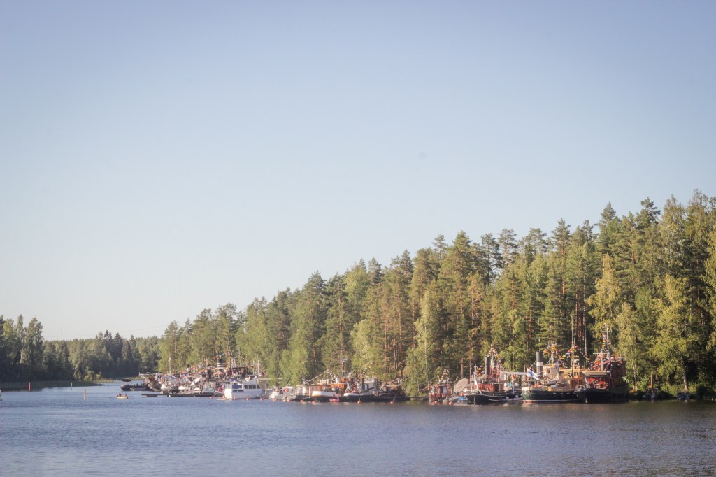 Kuvassa aurinkoinen järvimaisema. Paljon metsää ja höyrylaivoja.