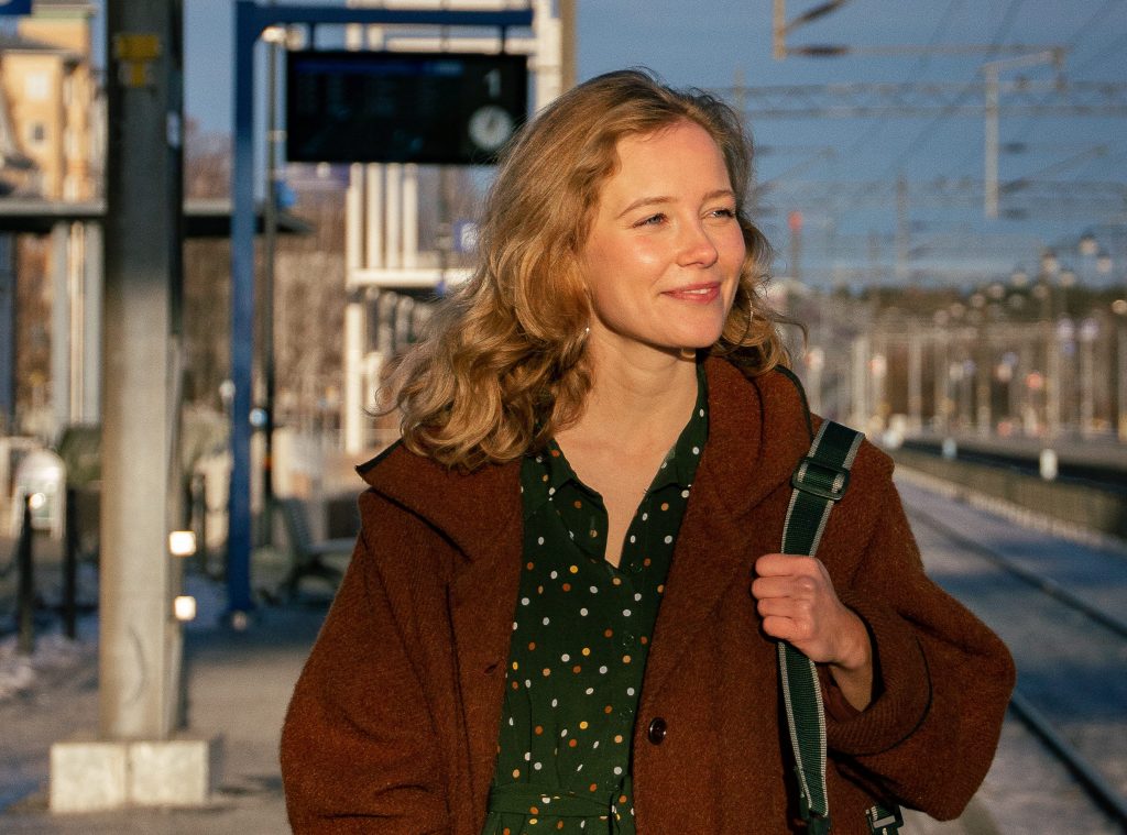 Lotta seisoo rautatieasemalla ruskeassa takissa ja vihreässä, pilkullisessa haalarissa. Takana näkyvät junaraiteet ja aikataulu. Lotta katsoo vasemmalle ja hymyilee.