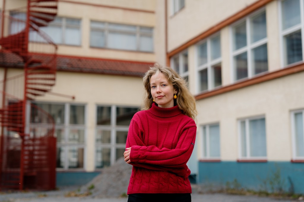 Lotalla on yllään punainen villapaita. Hän katsoo kameraan vakavana, kädet puuskassa. Takana näkyy koulurakennus.